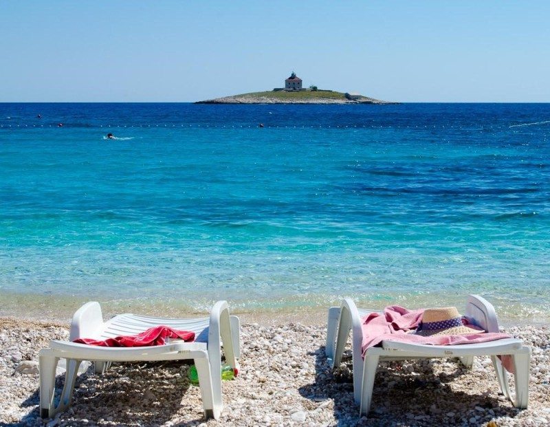 Duas espreguiçadeiras vazias na praia de Pokonji Dol, em Hvar, de seixos e virada para o mar azul-claro, com uma pequena ilha e um edifício ao fundo.