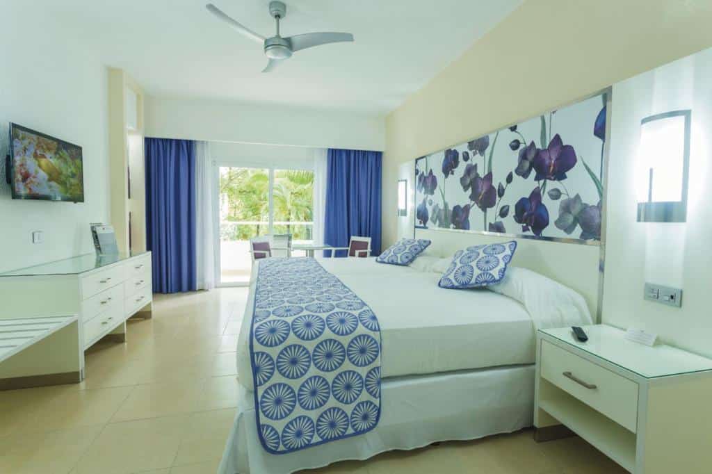 Quarto do Riu Playacar com cama de casal, decoração moderna e vista para a área exterior pela varanda.