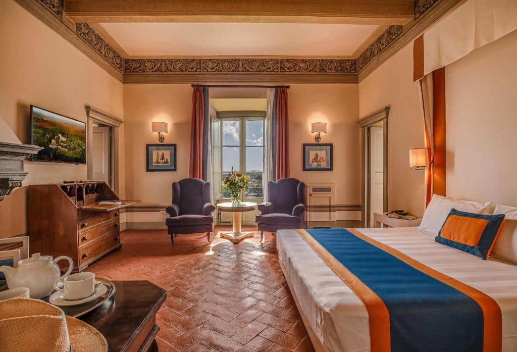 Suíte executiva do Borgo Scopeto Wine &amp; Country Relais, um dos hotéis na Toscana, com 55 m², cama grande de casal, duas poltronas roxas, mesa redonda ao centro com uma flor, janela a refletir a luz do sol e o céu azul com algumas nuvens, e móveis de época pelo quarto, com chão de tacos antigo