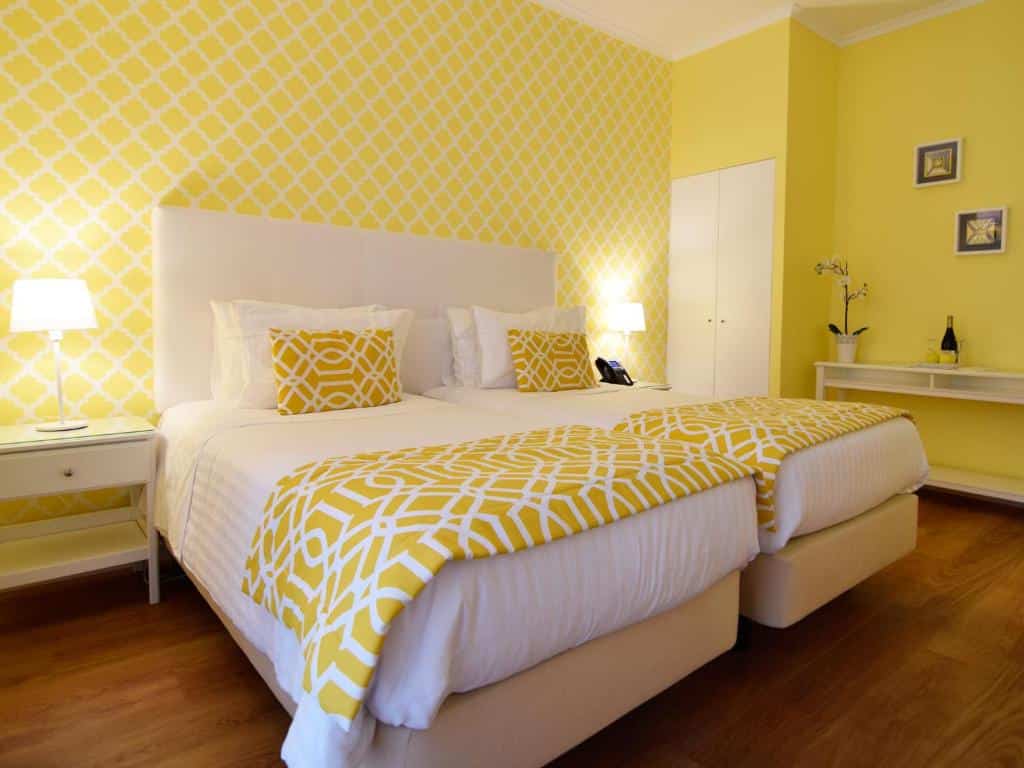 Quarto do Dream Chiado Apartments com duas camas de solteiro com almofadas, um pequeno roupeiro de duas portas e um aparador com um vaso de flores e uma garrafa de vinho, com decoração amarela e branca