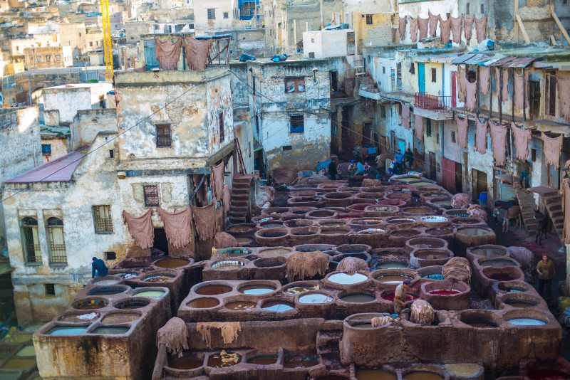 Vista de uma curtumaria com grandes tanques de tingimento rodeados de prédios antigos, numa zona urbana densa, com couro estendido a secar em Fez, Marrocos.
