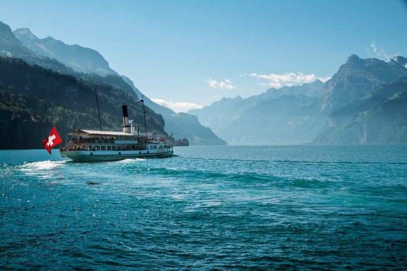 Um barco a navegar num rio na Suíça durante o dia, com montanhas ao fundo.