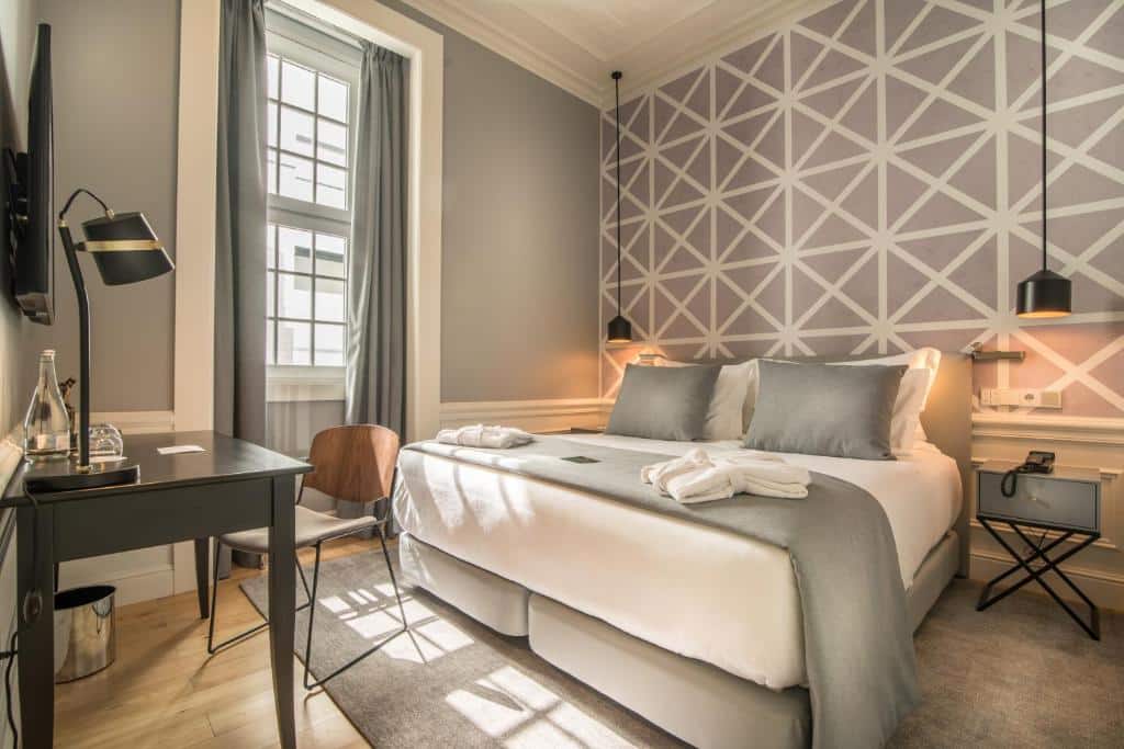 Quarto do Hotel da Baixa com cama de casal, secretária, cadeira, janela e uma parede com detalhes geométricos em tons neutros.