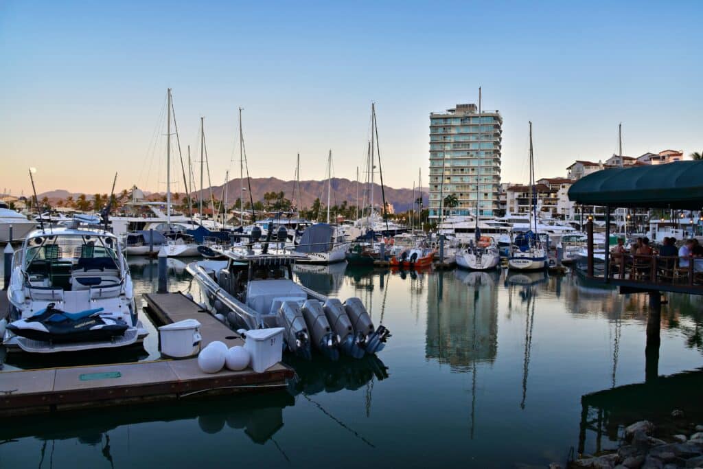 Barcos atracados numa marina com um edifício alto, montanhas e pessoas sentadas num restaurante ao ar livre ao fundo.