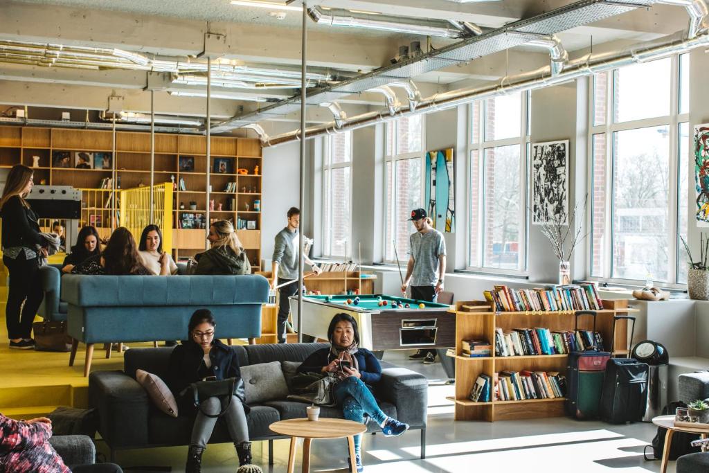 Pessoas sentadas em sofás a ler ou a usar dispositivos num lounge bem iluminado do ClinkNOORD Hostel, enquanto outras jogam matraquilhos junto a estantes de livros e janelas grandes.