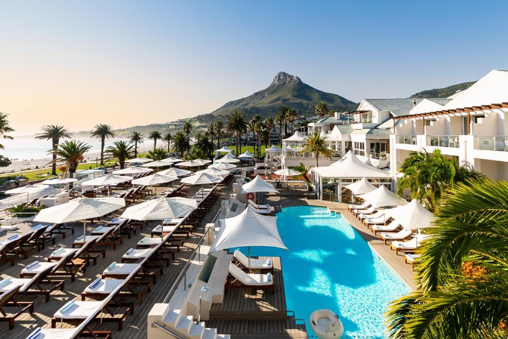 Zona de resort no The Bay Hotel com piscina, filas de espreguiçadeiras, chapéus de sol brancos, palmeiras e uma montanha ao fundo.