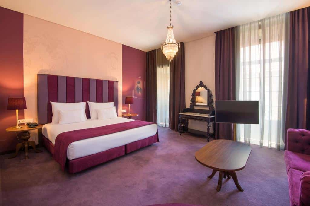 Quarto do Vila Galé Collection Braga com cama king-size, decoração em tons de roxo, candeeiro de teto, secretária com espelho e janelas amplas com cortinas.