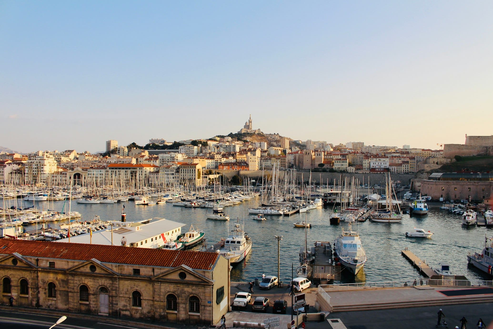Marina de Marselha repleta de barcos, com a cidade e uma igreja no topo da colina ao fundo sob céu azul.