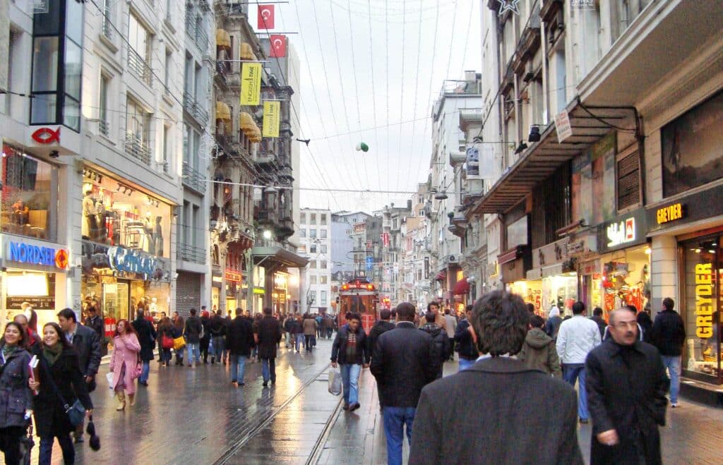 Uma rua movimentada com pessoas a caminhar, lojas de ambos os lados e um elétrico ao centro, num dia nublado.