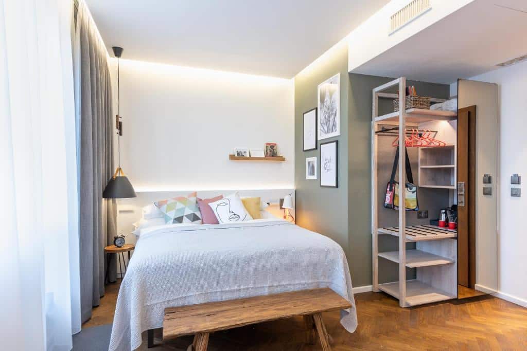 Quarto moderno do Mosaic Design Hotel, com cama de casal, roupeiro aberto, banco de madeira, arte na parede e luz natural suave.