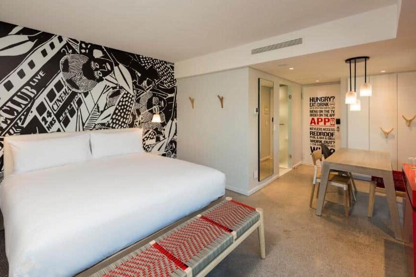 Quarto do Radisson RED Hotel com cama grande, arte gráfica na parede, mesa de refeições e casa de banho com paredes de vidro ao fundo.