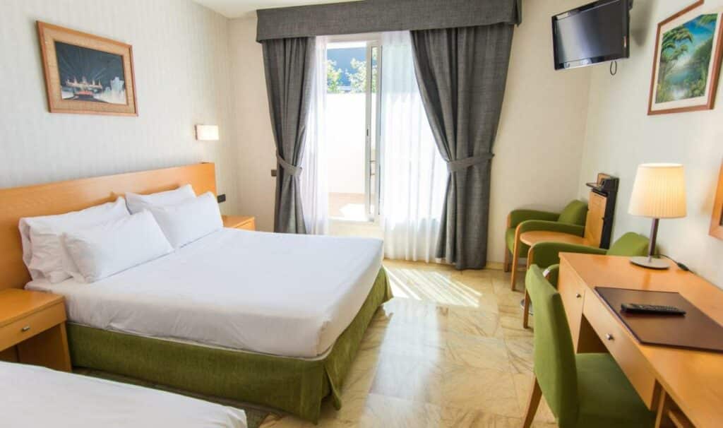 Quarto do Flor Parks, com cama de casal, secretária com cadeira, duas poltronas com mesinha e uma janela com cortinas cinzentas e brancas.