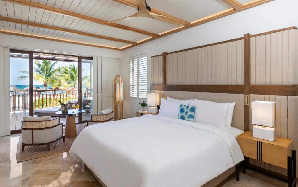 Quarto do Fairmont Mayakoba com cama king size, varanda e vista para praia tropical com coqueiros. Junto à varanda há poltronas.