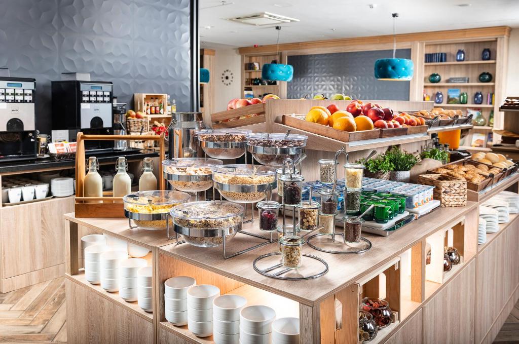 Um buffet de pequeno-almoço do Three Corners Lifestyle Hotel com cereais, fruta, doces, leite, chávenas e condimentos dispostos em prateleiras e balcões de madeira.