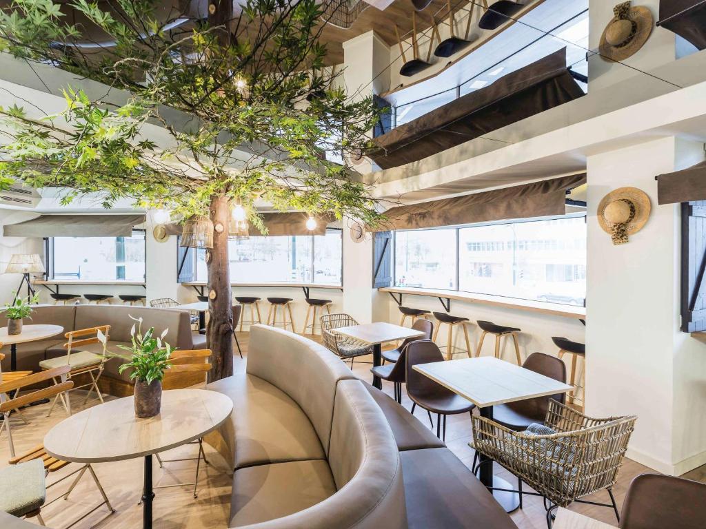 Área de convívio do Hotel Ibis Styles Lisboa Marquês de Pombal com várias janelas, mesas e cadeiras, alguns sofás e, ao centro, uma pequena árvore iluminada