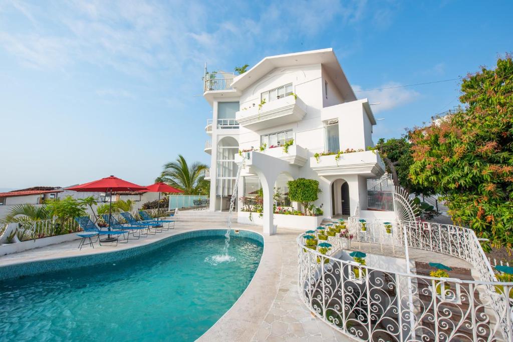 Uma casa branca de três andares com varandas, uma piscina curva, espreguiçadeiras, chapéus de sol vermelhos e plantas tropicais no logradouro.