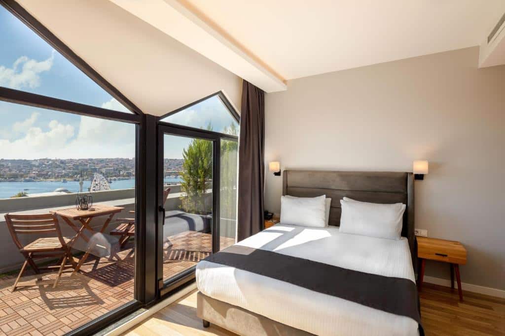 Quarto do Doruk Palas Hotel moderno com cama de casal, janelas grandes e varanda com vista para a água e a paisagem urbana ao longe.