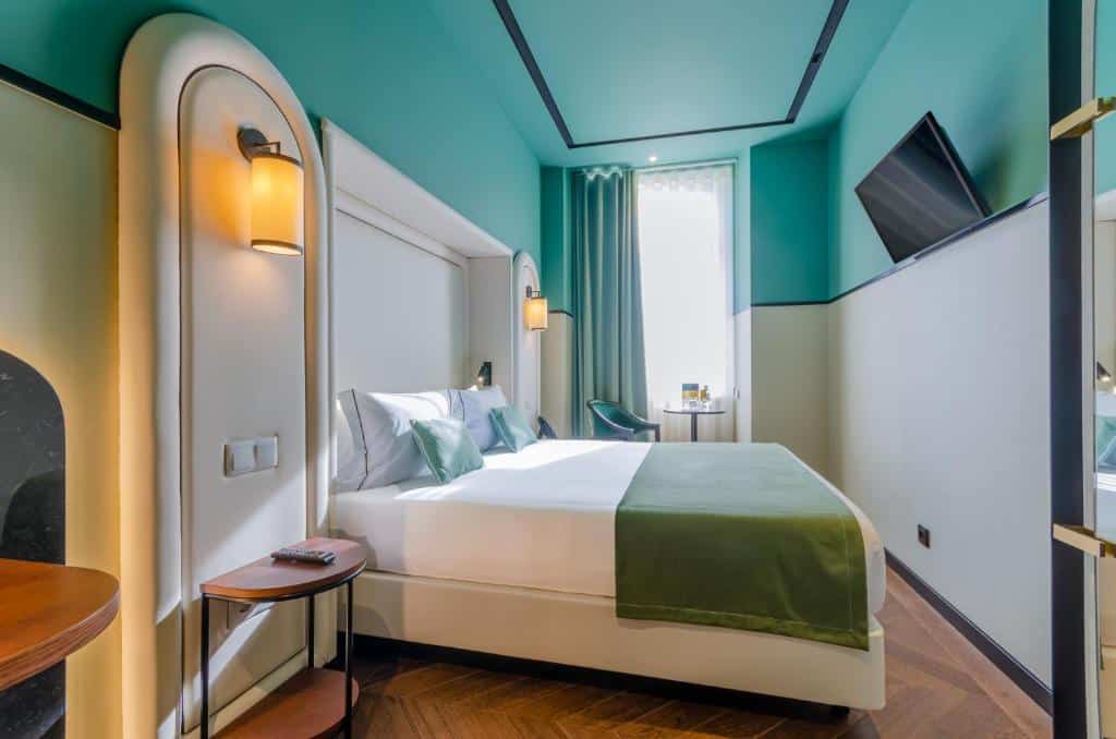 Quarto do Nicola Rossio Hotel com uma cama de casal, uma janela com cortinas, uma mesinha com uma cadeira estofada e, em frente à cama, uma televisão e uma mesa de cabeceira com um candeeiro de parede