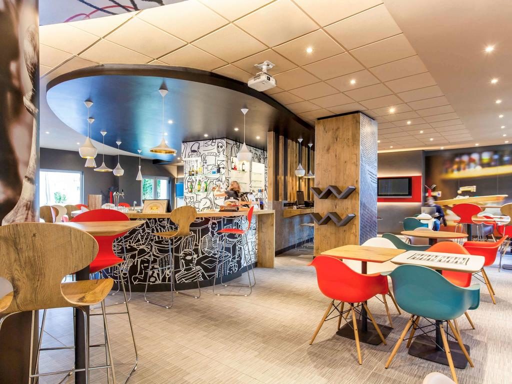 Área do restaurante do Ibis José Malhoa com mesas de madeira, cadeiras coloridas, uma televisão e um balcão de pedidos com bancos altos