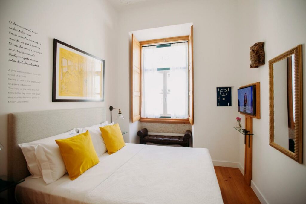 Quarto de casal no Otília Apartamentos, com almofada e quadro amarelos na parede, espelho em frente à cama e uma janela ampla ao fundo.