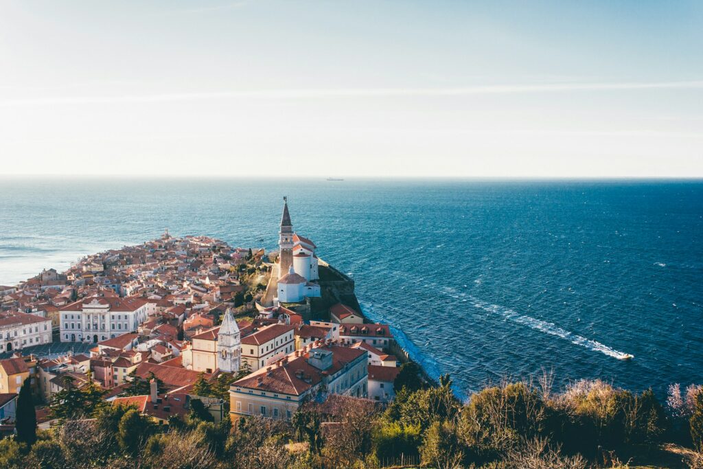 Vista aérea de Piran, cidade costeira na Eslovénia. A cidade é caracterizada por edifícios de telhados de telha e uma igreja com torre sineira pontiaguda. Situada numa península, é rodeada pelo azul brilhante do Mar Adriático. A imagem capta um barco a deixar um rasto na água, além da natureza e da arquitetura em harmonia.