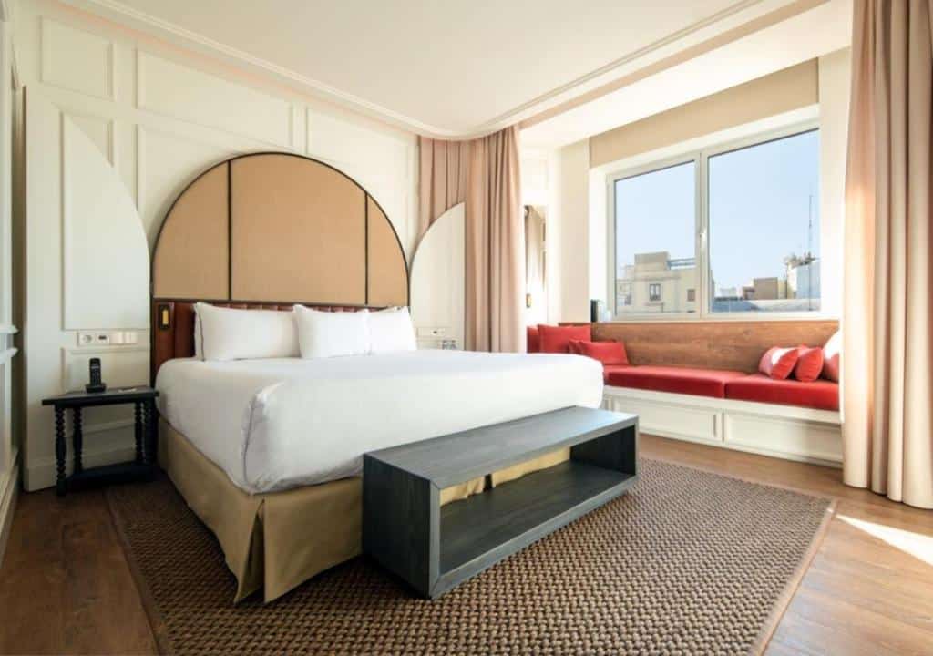 Quarto do Only YOU Hotel Valencia, com cama de casal, almofadas vermelhas, sofá e vista da cidade através da janela.