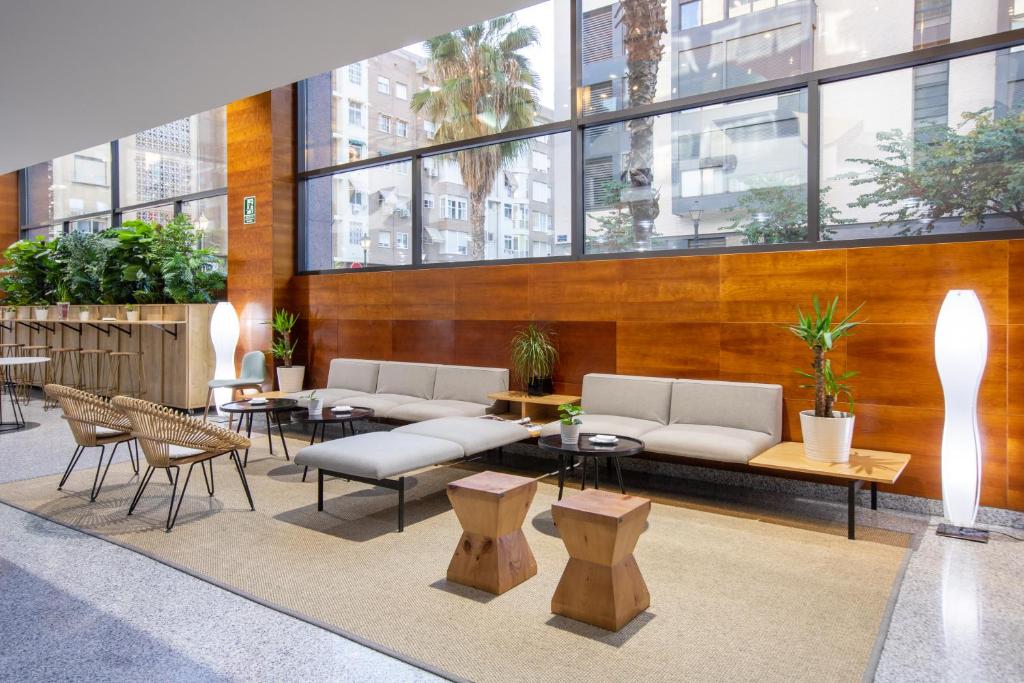 Sala de estar confortável do Eurostars Acteón, com sofás e poltronas em tons claros, mesa de centro de madeira e plantas, e amplas janelas com vista para edifícios e palmeiras.