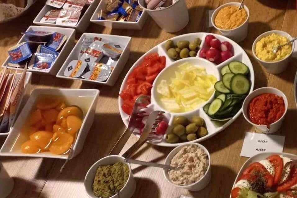 Uma mesa do Váralja Home com vários itens de pequeno-almoço, incluindo legumes fatiados, azeitonas, pastas, queijos e pêssegos em calda.