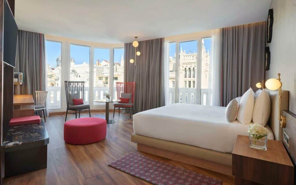 Quarto do Hyatt Centric Gran Via Madrid, um dos melhores hotéis em Madrid. A cama tem roupa de cama branca e mesinha de cabeceira com candeeiro dos dois lados. Há uma zona de estar com mesinha, cadeiras e pufe em frente à cama, e as paredes do quarto têm janelas com cortinas e vista para a cidade. Ao lado da zona de estar há uma secretária com cadeira e uma TV virada para a cama.