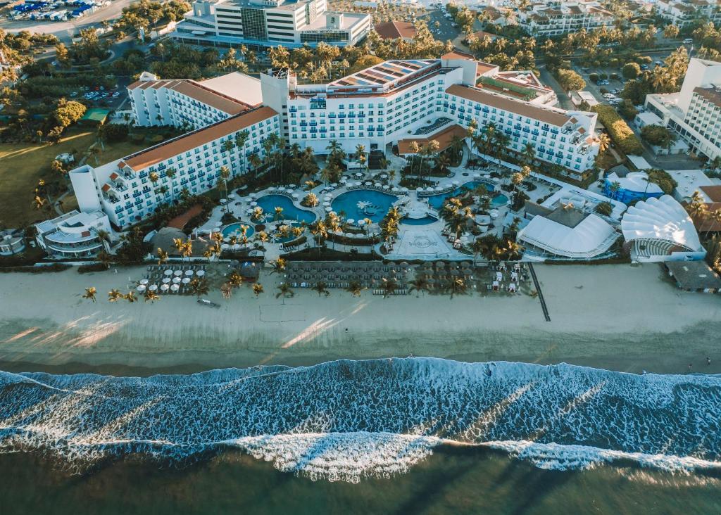 Vista aérea de um grande resort à beira-mar com piscinas, palmeiras e espreguiçadeiras ao longo de uma costa arenosa próxima ao oceano.
