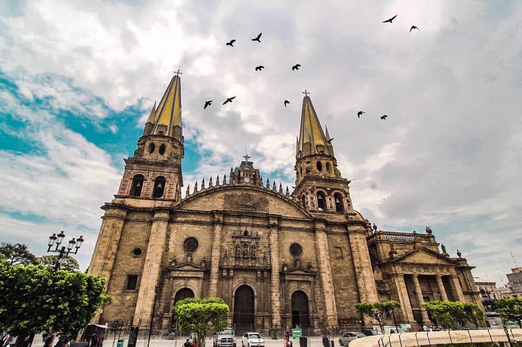 Catedral de Guadalajara, grande edifício religioso de arquitetura colonial espanhola com torres gémeas douradas num céu nublado, com bandos de pássaros a voar.