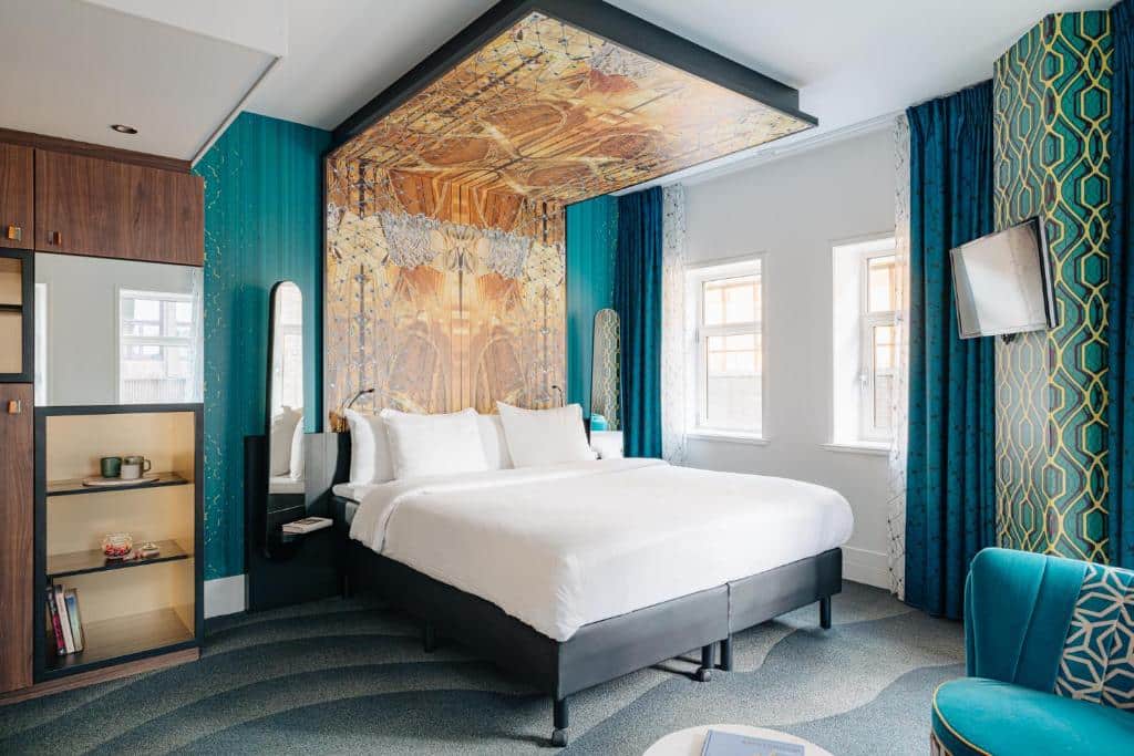 Quarto do Eden Hotel Amsterdam, que ilustra onde ficar em Amesterdão: cama grande, decoração em azul-petróleo e dourado, arte na parede por cima da cama, duas janelas e uma TV na parede.