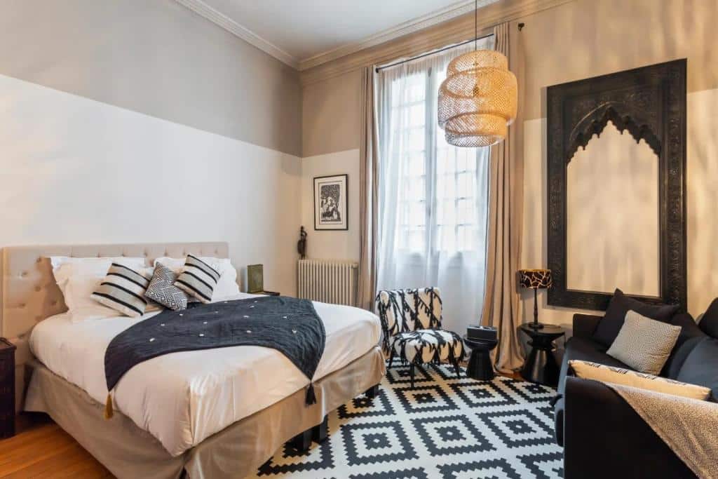 Quarto moderno do Sourire Boutique Hôtel Particulier com cama de casal, tapete geométrico, poltrona e uma grande janela com cortinas.