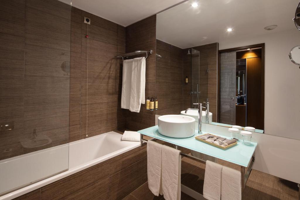 Casa de banho do NEYA Lisboa Hotel com banheira do lado esquerdo, um espelho amplo por cima do lavatório de cuba branca, vários produtos de higiene e toalhas brancas.