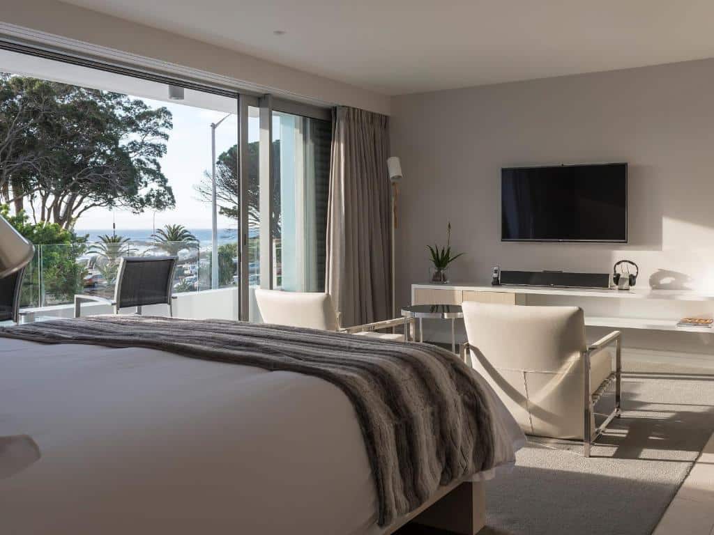 Quarto do South Beach Camps Bay Boutique Hotel com cama grande, duas cadeiras, TV na parede e varanda com vista para o mar e árvores.
