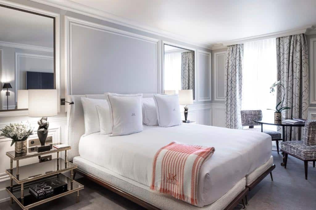 Cama com roupa branca no J.K. Place Paris, com duas mesas de cabeceira com candeeiros e uma secretária num quarto moderno e luminoso.