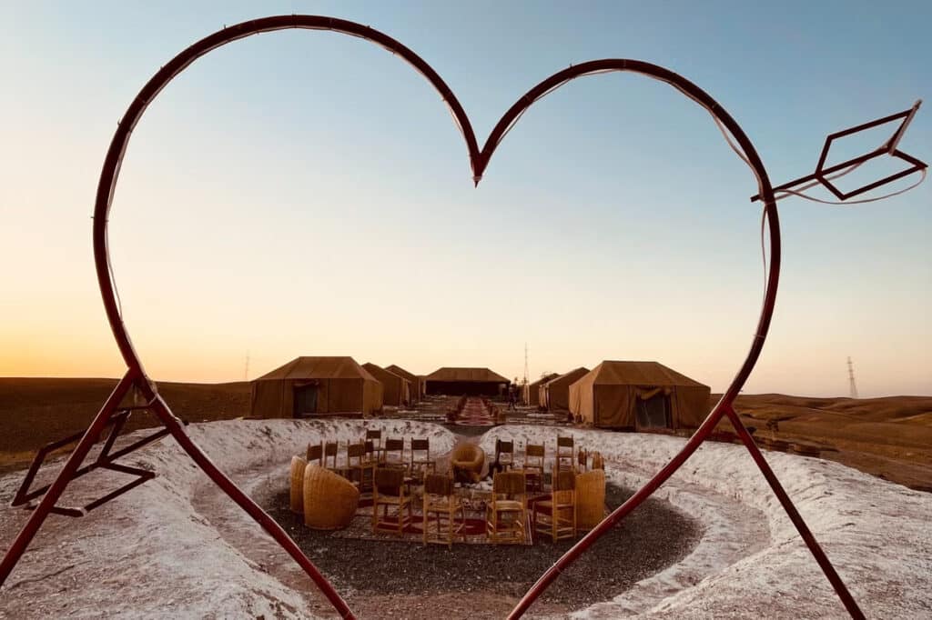 Escultura em forma de coração emoldura um acampamento no Deserto de Agafay, com tendas castanhas e uma área central com cadeiras e mesas ao pôr do sol.