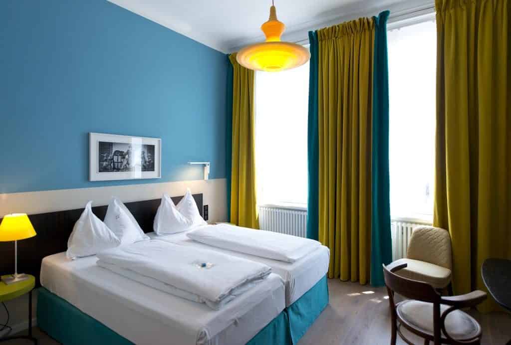 Quarto no Hotel Beethoven, com cama de casal, paredes azuis, cortinados amarelos e uma secretária com duas cadeiras junto a grandes janelas.