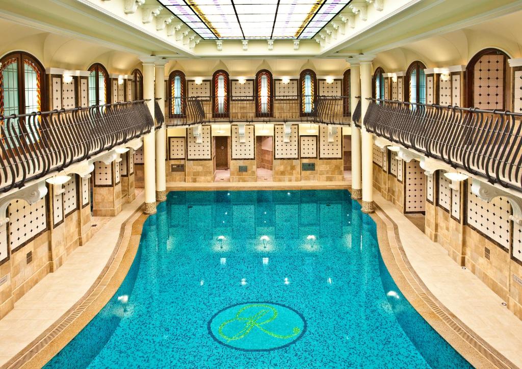 Piscina interior do Corinthia Budapest, com águas claras, rodeada por janelas em arco, varandas e teto de vidro.