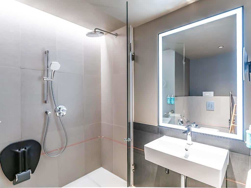 Casa de banho moderna com chuveiro, lavatório suspenso, espelho grande e azulejos cinza neutros.