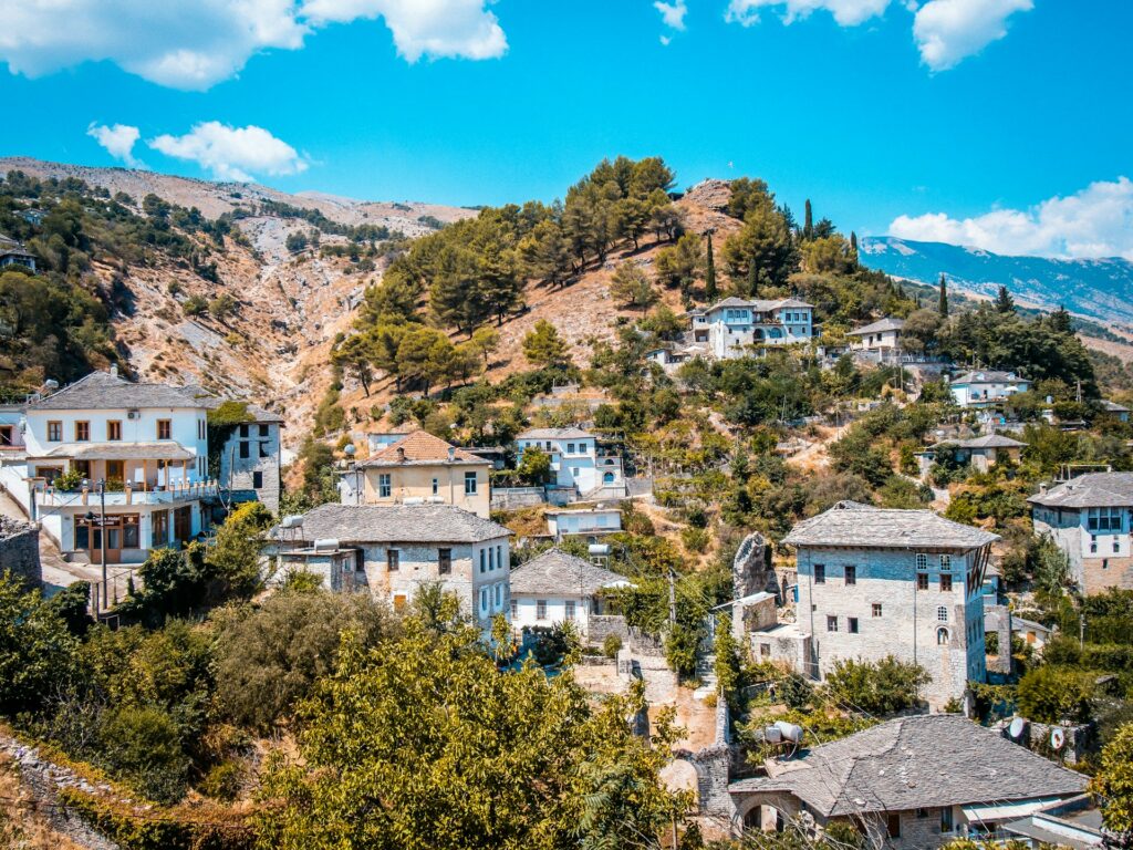 Uma aldeia de Gjirokastër, um dos destinos invulgares da Europa, encravada em encostas montanhosas, com casas de pedra de telhados cinzentos. A paisagem é pontuada por árvores e arbustos verdes, e as montanhas ao fundo estão parcialmente cobertas por vegetação rasteira. O céu está limpo, com algumas nuvens esparsas.