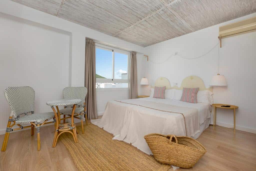 Quarto bem iluminado da Can Beia Hostal Boutique com cama de casal, duas mesas de cabeceira, um cesto de vime e uma pequena mesa com duas cadeiras junto à janela.