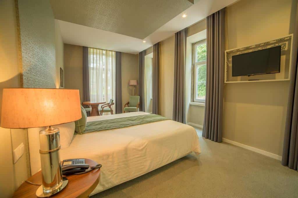 Quarto do Hotel do Parque com cama de casal, mesa de cabeceira, candeeiro, TV de parede, cadeiras e janelas amplas com cortinas.