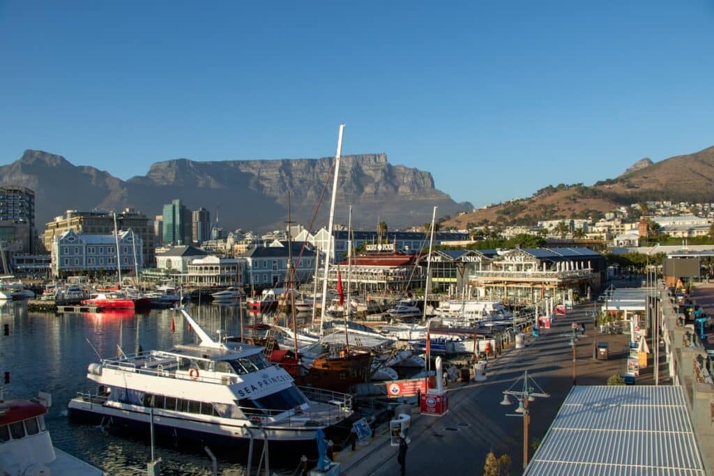 Porto do V&A Waterfront com barcos atracados, edifícios à beira-mar e a Table Mountain ao fundo num céu azul claro.