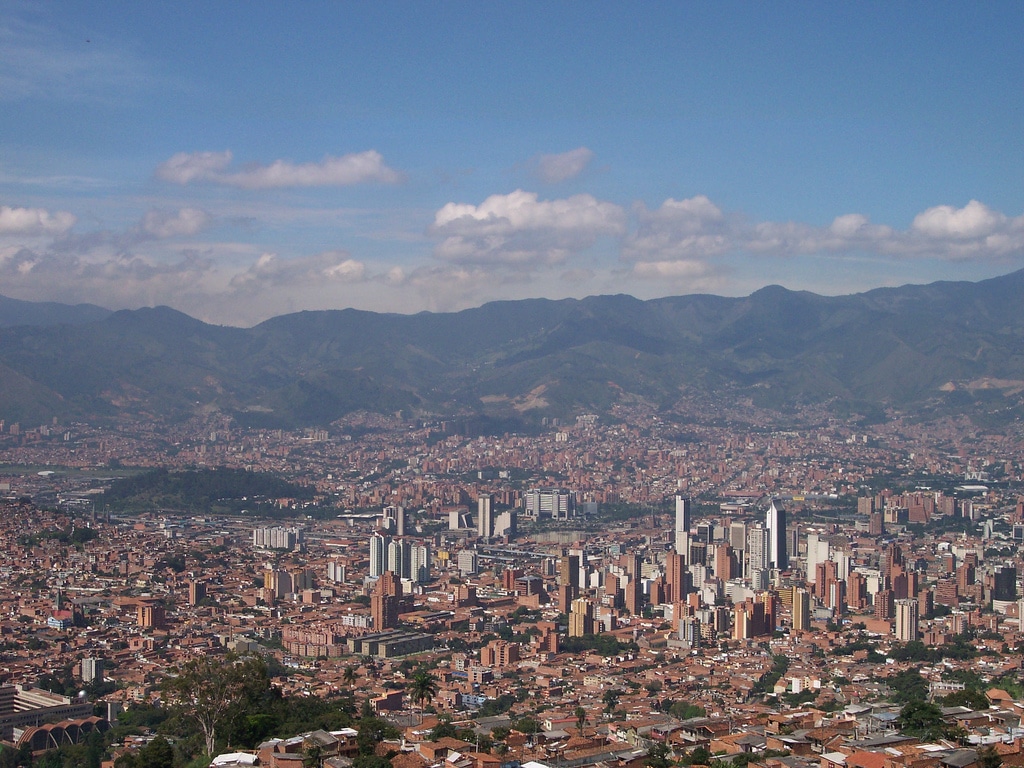 Vista panorâmica de Medellín, com uma mistura de edifícios altos e zonas residenciais, tendo ao fundo montanhas verdes sob um céu azul.