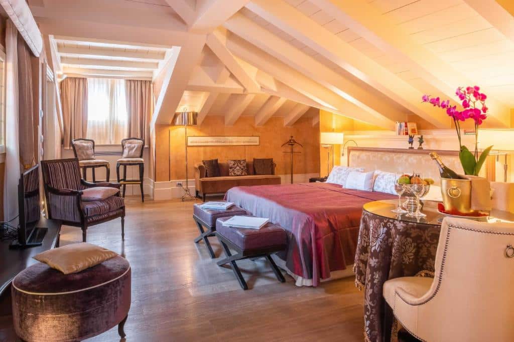 Quarto de hotel luxuoso com teto alto e vigas à vista, decorado em tons de vermelho, castanho e dourado, com edredão vermelho-vinho na cama de casal e dois sofás de couro junto à janela