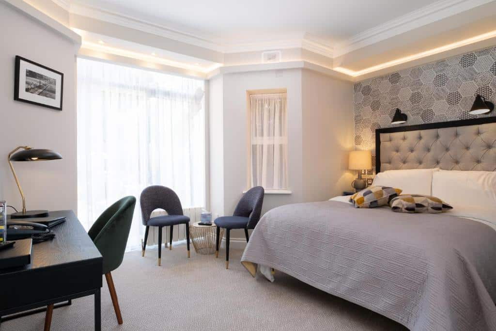 Quarto no Roxford Lodge Hotel com cama king size, cabeceira estofada cinzenta, duas poltronas azuis e secretária preta moderna. Iluminação ambiente e vista para o exterior por janelas amplas.