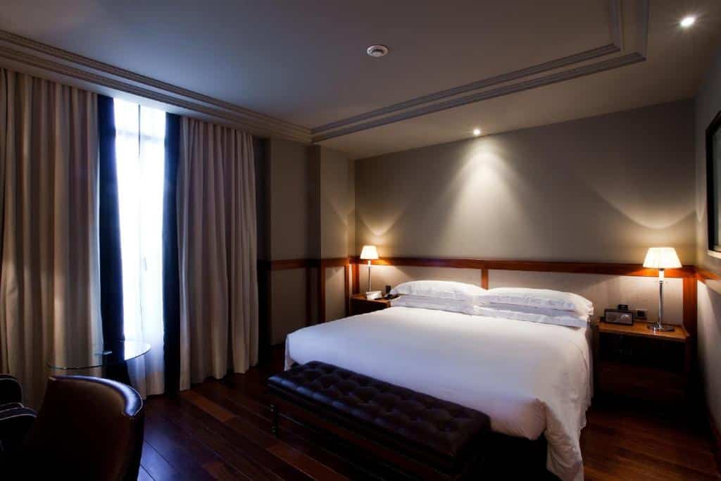Quarto do Hotel 1898, com cama de lençóis brancos, candeeiros de cabeceira de ambos os lados e um banco de couro castanho aos pés. Junto à janela, com cortinas bege, há uma secretária com cadeira.