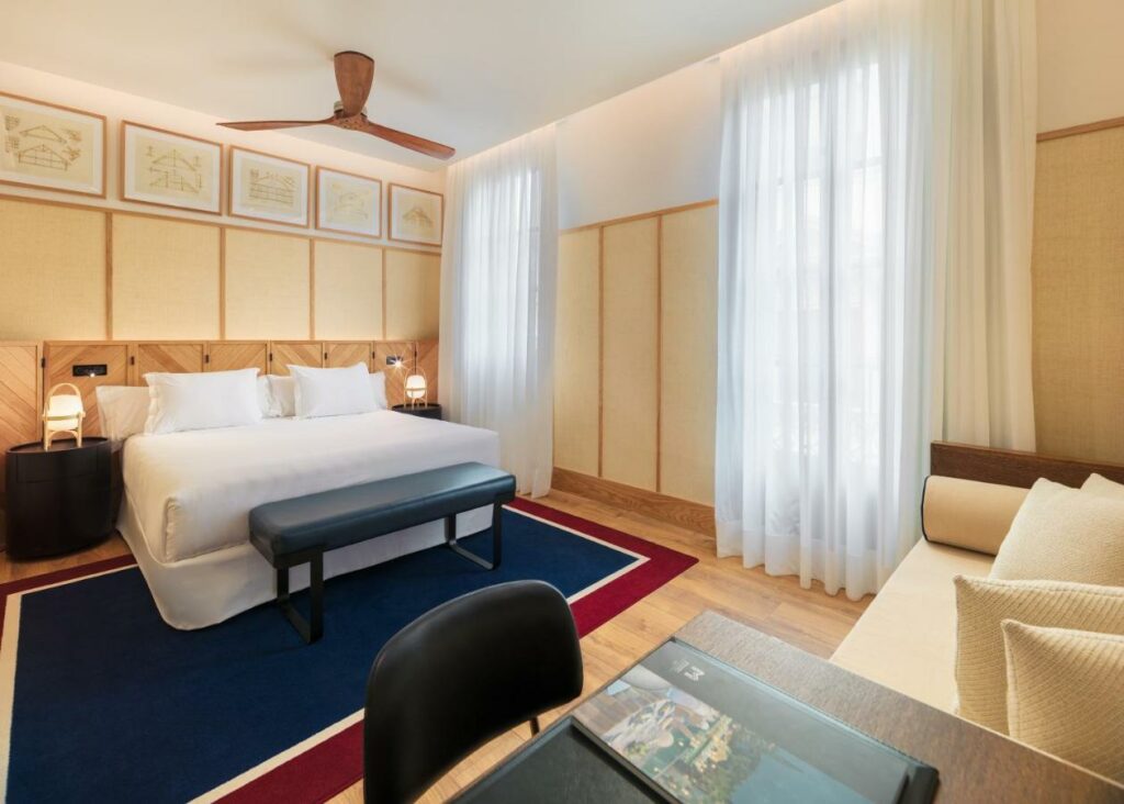 Quarto do H10 Madison. Uma cama de casal com mesas de cabeceira e candeeiros, ventoinha de teto, sofá e secretária junto a duas janelas com cortinas brancas.