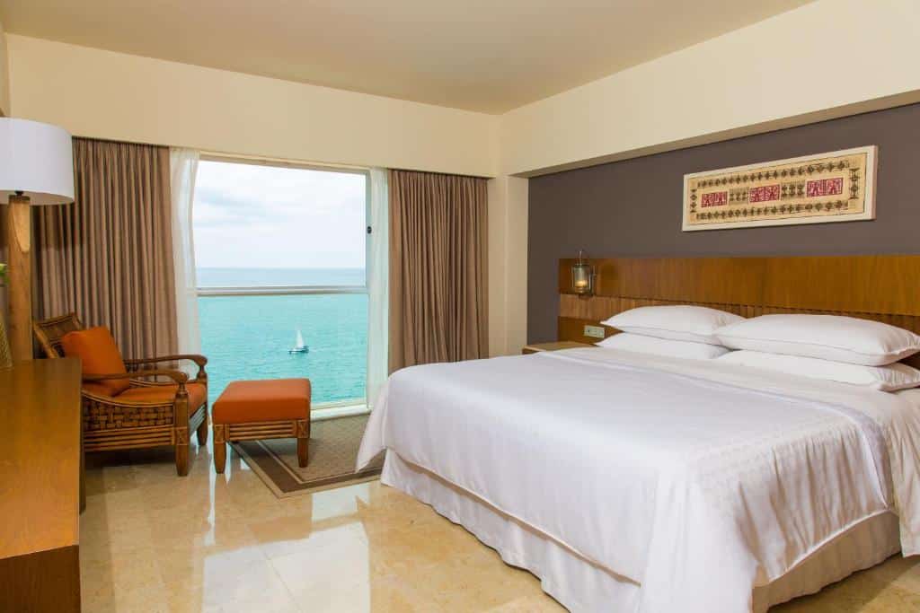 Quarto do Sheraton Buganvilias Resort & Convention Center com uma cama grande, duas cadeiras junto a uma janela e vista para o oceano e um veleiro.